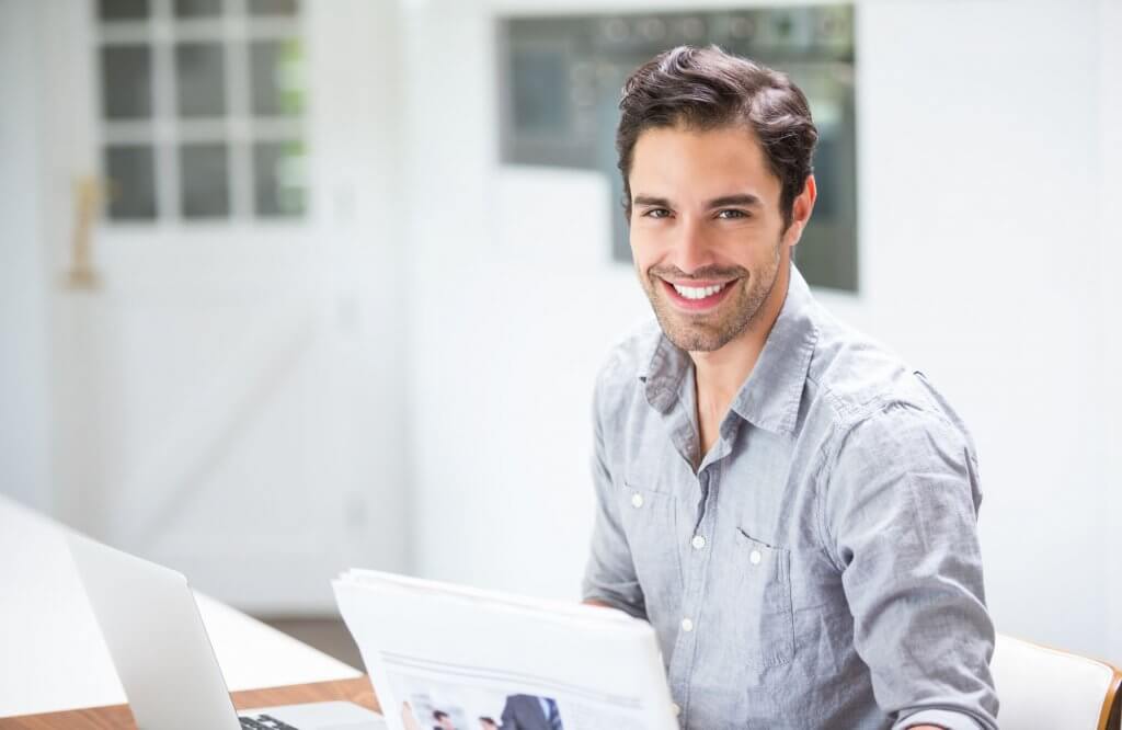 Portrait Of Smiling Young Man Holding Documents While Sitting At Desk With Laptop At Home